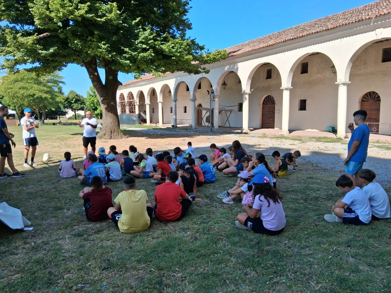 Un gruppo di bambini seduti sul prato davanti a un porticato, durante un centro estivo Lilliput